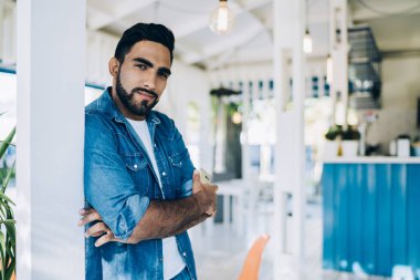 Kollarını çapraz bağlamış, elinde telefonla yakışıklı etnik bir adam ara verip sokak kafesinde kameralara bakıyor.