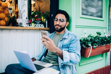 Kot pantolon ve gözlüklü, şehir binasının yanında dizüstü bilgisayarı olan ve cep telefonuyla test uygulaması kullanan pozitif sakallı bir erkek. 