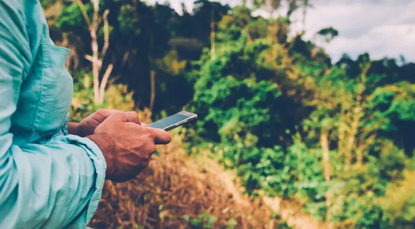 Side view of male traveler holding mobile phone in hands and reading message via application during travel to National Park.Tourist man using smartphone outdoors. Online navigation with cellphone