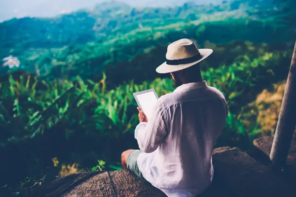 Young male tourist digital nomad working online on touch pad during summer vacations.Man wanderlust reading book on tablet while sitting outdoors inspired by scenery Amazonian nature.Copy scape screen