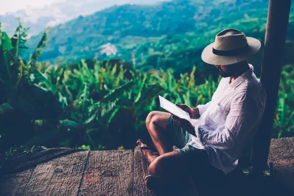 Young male tourist digital nomad working online on touch pad during summer vacations.Man wanderlust reading book on tablet while sitting outdoors inspired by scenery Amazonian nature.Copy scape screen