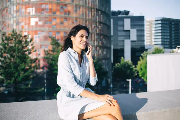 Cheerful millennial woman in casual wear talking on mobile phone laughing spending time on urban setting background, smiling female making smartphone call in roaming resting on weekend in trip
