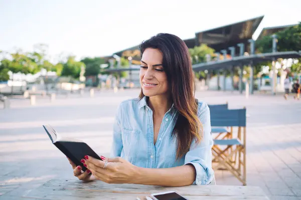 Intelligent positive woman in smart casual wear reading interesting book novel outdoors on sunny day, smiling 30s female brunette satisfied with free time spending on literature hobby on cafe terrace