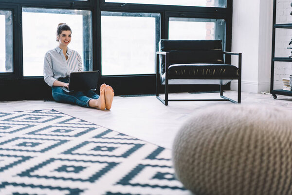 Cheerful young woman smiling while updating software on modern laptop for remote work sitting on floor in apartment with stylish interior, happy Caucasian freelancer using wireless home internet