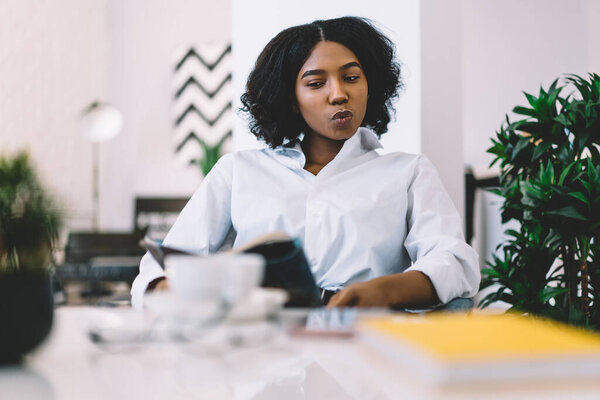 African American hipster girl with dark skin reading fresh publication from newspaper interested with fashion news, black female youngster enjoying home recreation weekend with education literature