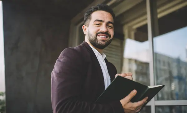 Low angle of happy ethnic male in formal wear smiling and looking at camera while writing in notepad outside contemporary building