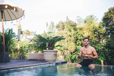 Çıplak gövdeli ve siyah yüzme şortlu genç yakışıklı erkek Bali spa merkezindeki tropikal bitkiler arasında yüzme havuzunun kenarında oturup yemek yiyor.