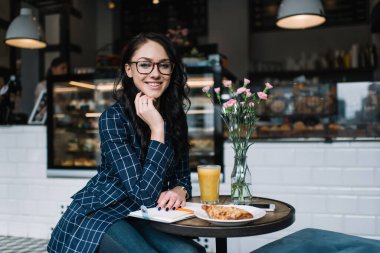 Gözlüklü neşeli bayan uzaktan kumandalı çalışanı kafeteryada müşteriyle buluşmayı beklerken uzaktan çalışıyor ve kameraya bakıyor.