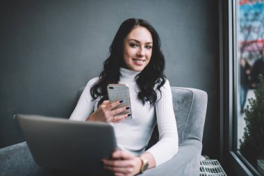 Beyaz kazaklı mutlu kadın serbest yazar rahat koltukta oturuyor ve modern kafe molasında akıllı telefonlara bakarken gülümsüyor.