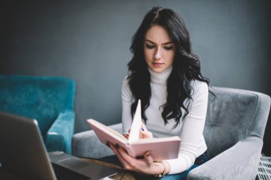 Evdeki modern oturma odasındaki dizüstü bilgisayarlı koltukta otururken gündelik kıyafetlerle gezen düşünceli genç bir bayan.