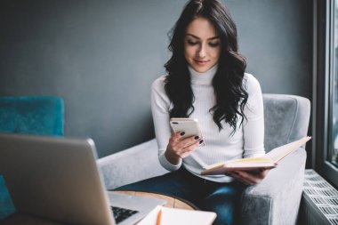 Sırıtkan, gündelik giysiler içinde, masadaki pencerenin kenarına dizüstü bilgisayar ve defterle oturmuş, açık bir kitap tutarken ve evde akıllı telefon kullanırken gülümseyen genç bir kadın.