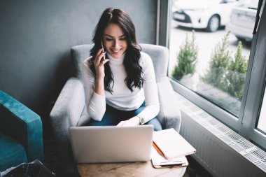 Yaratıcı iş yerinde oturan, cep telefonuyla konuşan, dizüstü bilgisayar kullanan ve iş projesinde uzaktan çalışan pozitif bir kadın.