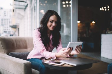 Genç bayan serbest çalışan masadaki dizüstü bilgisayarlı ve akıllı telefonlu koltukta otururken çağdaş kafeteryadaki uzaktan çalışma sırasında gülümseyerek ve tabletle video görüşmesi yapıyor.