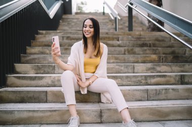 Moda kıyafetleri içinde neşeli, genç bir bayan merdivenlerde oturuyor ve gün ışığında selfie çekmek için cep telefonu kullanıyor.