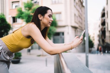 Sarı tişörtlü, korkuluklara yaslanan ve şehir manzarasının fotoğrafını çeken genç bayanın yan görüntüsü.