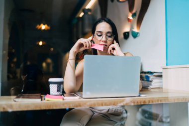 Gözlüklü, laptopta çalışırken yapışkan not ısıran ve çalışma odasında bir fincan kahveyle oturan konsantre genç bir kadın.