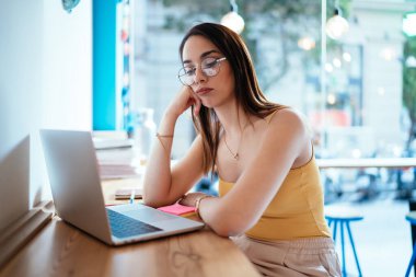 Gündelik kıyafetleri ve gözlükleri olan üzgün genç bir kadın internetten bilgisayarını kullanarak duvarın yanındaki kafe masasında oturuyor.