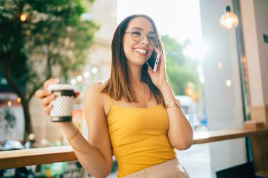 Gülümseyen esmer kadın gündelik kıyafetler ve gözlüklerle elinde bir fincan sıcak kahve tutarken çağdaş kahve dükkanında akıllı telefonla konuşuyor.