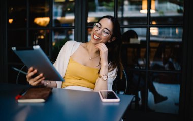 Gözlüklü, günlük kıyafetleri olan, elinde tablet olan, modern sokak kafesindeki masada oturan, gülen genç bir kadın serbest yazar.