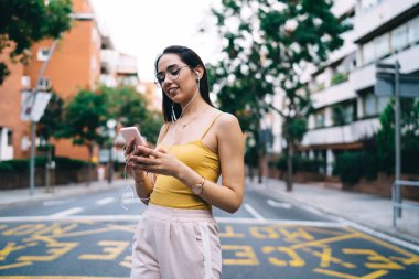 Moda gözlükleri takmış, kulaklıklı, cep telefonuyla mesajlaşan ve müzik dinleyen genç bir kadın.