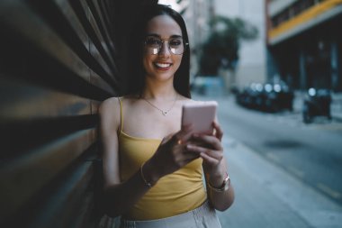 Pozitif genç bayan kameraya bakıyor ve elinde akıllı telefonu tutarken gülümsüyor. Bu arada telefon akşam caddesinde yüzünü aydınlatıyor.