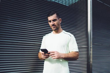 Tişörtlü, ciddi sakallı bir adam. Güneşli bir günde pencerenin yanında duruyor. Jaluzi ve akıllı telefonuyla kameraya bakıyor.