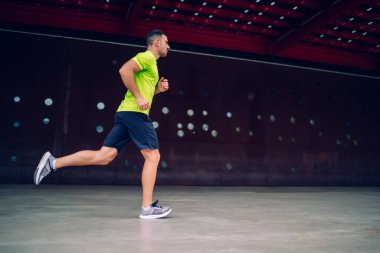 Aktif giyimli ve spor ayakkabılı güçlü erkek koşucunun yan görüntüsü şehrin modern binasının duvarının yanındaki boş kaldırımda hızla koşuyor.