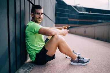 Acımasız yetişkin ciddi sporcunun yan görüntüsü asfaltta oturuyor, cep telefonuna bakarken ve kameraya bakarken dinleniyor.