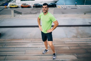 Spor kıyafetleri ve spor ayakkabıları giyen, merdivenlerin ortasında durup gülümseyen ve kalçalarına dokunan pozitif bir sporcu.