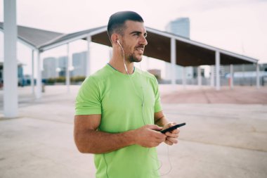 Günlük kıyafetleri ve kulaklıkları olan genç bir erkeğin sokak koşusuna hazırlanırken müzik dinlediği ve akıllı telefonuna göz attığı yan görüntü.