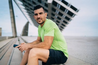 Spor kıyafetli, taş merdivenlerde oturan ve dışarıda idmandan sonra dinlenirken kameraya dalgın dalgın bakan ciddi etnik erkek.