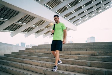 Aktif giyimli, spor ayakkabılı, spor ayakkabılı ve spor ayakkabılı, spor yaptıktan sonra geometrik güneş panelleriyle yapım aşamasında merdivenlerden inen, düşük açılı, olgun bir sporcu.