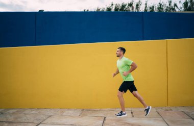 Şehir caddesindeki boş yolda sarı mavi duvarın yanında koşan aktif kıyafetli bir erkek koşucunun yan görüntüsü