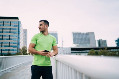 Kendine güvenen, spor kıyafetli, köprüde dikilmiş müzik dinleyen ve başka tarafa bakan kendine güvenen etnik sakallı bir erkekten.