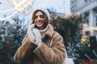 Kış kıyafetleri içinde genç bir kadın Noel süsleriyle sokakta duruyor ve eldiven takıyor.