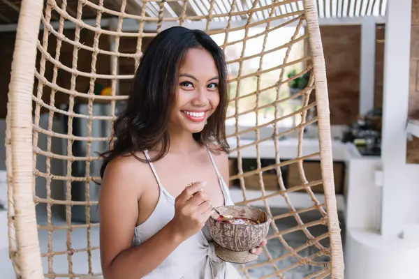 Delighted young ethnic female traveler smiling while sitting in wicker hanging egg chair and eating yummy breakfast in coconut bowl in Bali resort