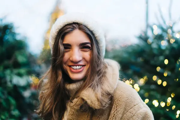 Smiling woman with long hair in hat and warm coat standing on background of Christmas trees in garlands and enjoying free time