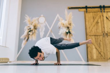 Spor kıyafetli bir adam vücudunu esnetirken ve yoga dersinde dengeyi geliştirirken Eka Pada Koundinyasana 'yı taklit ediyor.