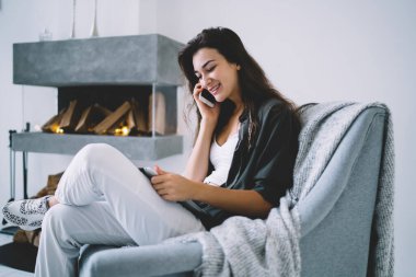 Rahat koltukta oturan ve kendini izole ederken akıllı telefondan konuşan sıradan giyinmiş genç bir kadının yan görüntüsü.