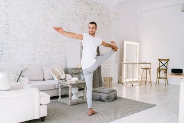 Spor kıyafetleri giymiş, çıplak ayaklı erkek. Ayaklarını esnetiyor ve evde el pençe divan duruyor.