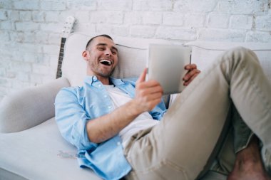 Genç pozitif sakallı erkek tablet üzerinde komik filmler izliyor ve evdeki oturma odasında rahat bir kanepede dinlenirken gülüyor.