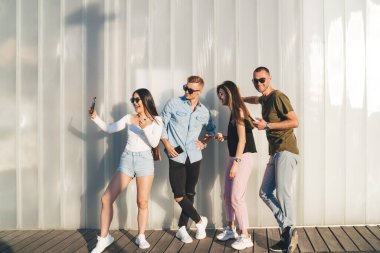 Gündüz kıyafetleri ve güneş gözlüğü takmış neşeli arkadaşlarla dolu bir vücut. Güneşli bir günde duvarın yanında durup selfie çekiyorlar.