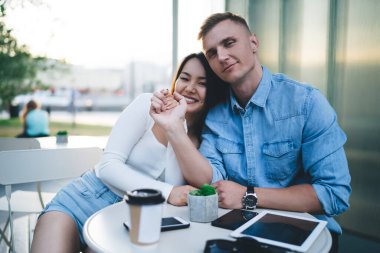 Yakışıklı, beyaz tenli, neşeli kız arkadaşına sarılan, hafta sonunu sokak kafeteryasında geçiren, kaldırım masasında kameraya gülümseyen, birbirine aşık genç bir çiftin yarım boy portresi.