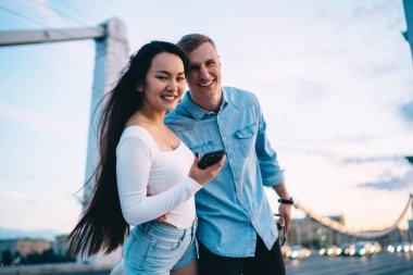 Şehirde yürürken kameraya gülümseyen neşeli bir çiftin yarım boy portresi. 20 yaşındaki neşeli hippi adamlar ellerinde akıllı telefon cihazları ve şehir köprüsünde poz verirken gülüyorlar.