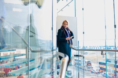 Sarışın, turkuaz ceketli genç ve güzel bir bayan işçi. Parlak ofis binasında merdivenlerden aşağı inerken kameraya bakıyor.