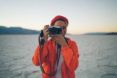 Kaliforniya 'da kuru çölde dururken sıcak kıyafetler içinde gülümseyen kadın fotoğrafçı retro fotoğraf makinesiyle fotoğraf çekiyor.