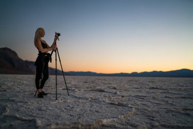 Yazlık kıyafetleriyle gezen kadın gezginin yan görüntüsü. Tripod 'un yanında durup Ölüm Vadisi' nin dağlık arazisinde gün batımının fotoğrafını çekiyor.