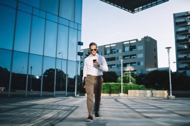 Moda giyinmiş genç işadamı, internet bankacılığı için akıllı telefon platformu üzerinden internet rezervasyonları yapıyor, yakışıklı erkek yönetici yürüyor ve finans haberleri için cep telefonuyla telsize bağlanıyor.