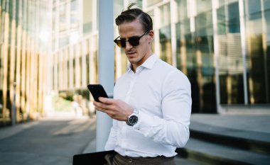 Moda giyinmiş genç işadamı, akıllı telefon platformu üzerinden banka adına internet rezervasyonları yapıyor. Güneş gözlüklü yakışıklı erkek yönetici, finansal haberleri aramak için cep telefonuyla telsize bağlanıyor.