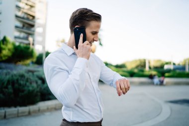 Kendinden emin erkek girişimci takılabilir kol saatinde buluşma zamanını kontrol ediyor. Detayları görüşmek için partneri arıyor. Genç işadamı cep telefonuyla aramak için gezici internet bağlantısını kullanıyor.